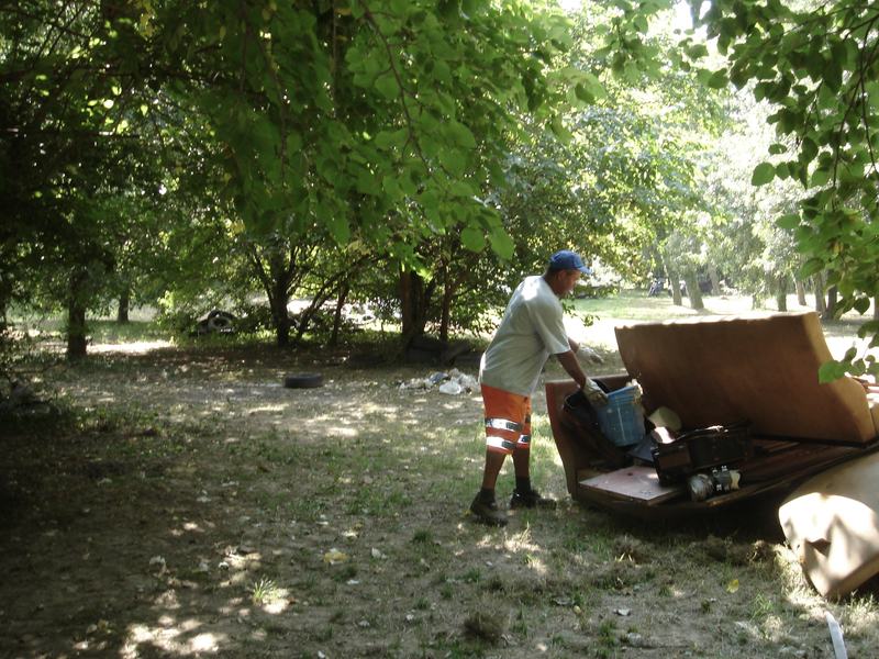Una persona con guanti da lavoro e pantaloni ad alta visibilit&agrave; carica rifiuti ingombranti tra gli alberi.